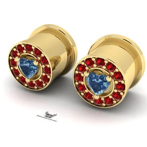 Puede incluir: Un par de tapones para los oídos dorados. Cada tapón presenta una piedra preciosa azul claro en forma de corazón en el centro, rodeada de pequeñas piedras preciosas rojas. La joya está sobre un fondo blanco.