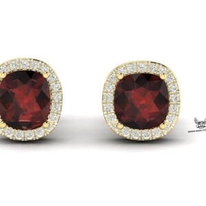 Peut inclure: Une paire de boucles d'oreilles à clous dorées avec des pierres précieuses rouge foncé taillées en coussin. Chaque boucle d'oreille est entourée d'un halo de petits diamants étincelants. Sur fond blanc.