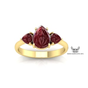 Puede incluir: Un anillo de oro con tres piedras preciosas rojas en forma de pera. La piedra central es más grande que las dos piedras a cada lado.