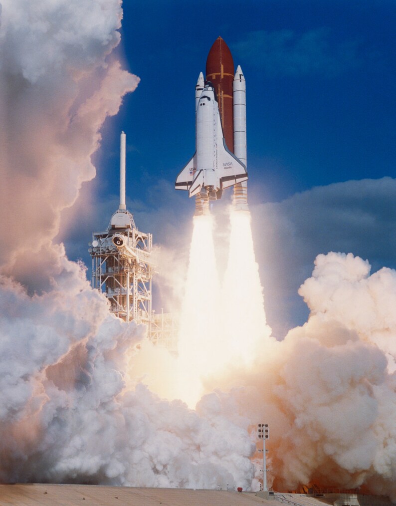 Op de afbeelding: Een witte en rode spaceshuttle lanceert de lucht in, en laat een spoor van witte rook en vuur achter. De shuttle wordt omringd door wolken.