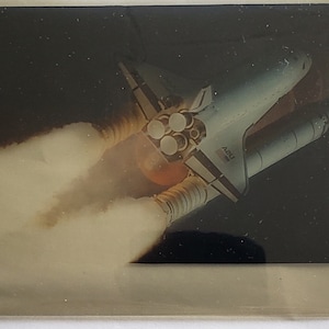 Op de afbeelding: Een witte spaceshuttle met de tekst "42B" aan de zijkant, die de ruimte in wordt gelanceerd met een spoor van rook en vuur.