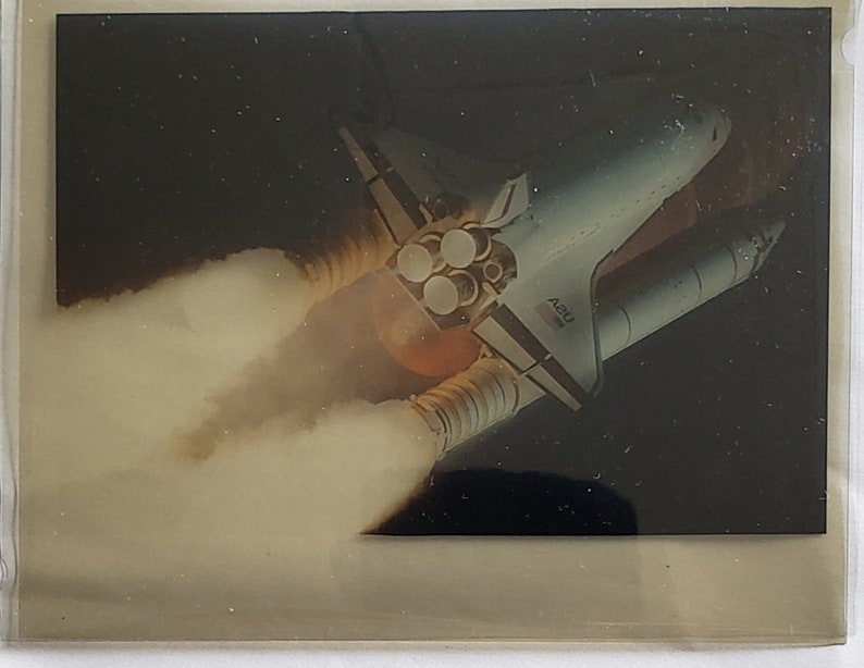 Op de afbeelding: Een witte spaceshuttle met de tekst "42B" aan de zijkant, die de ruimte in wordt gelanceerd met een spoor van rook en vuur.