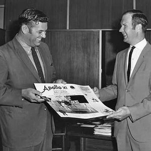 Op de afbeelding: Een zwart-witfoto van twee mannen in pak. De man links houdt een tijdschrift vast met de titel "Apollo XI" op de omslag. De man rechts glimlacht.