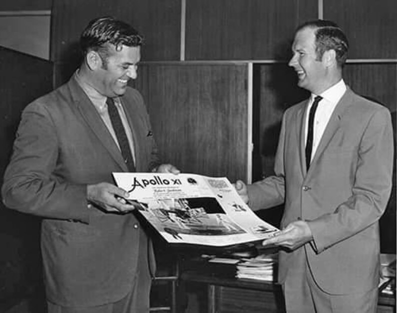 Op de afbeelding: Een zwart-witfoto van twee mannen in pak. De man links houdt een tijdschrift vast met de titel "Apollo XI" op de omslag. De man rechts glimlacht.