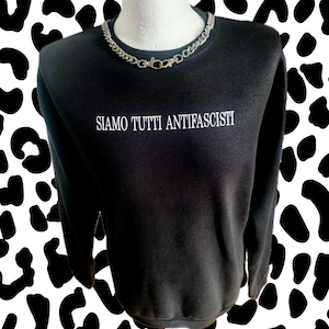 Könnte beinhalten: Schwarzes Sweatshirt mit dem weißen Text "SIAMO TUTTI ANTIFASCISTI". Eine silberne Halskette liegt um den Halsausschnitt. Der Hintergrund ist weiß mit schwarzen Flecken.
