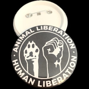 Könnte beinhalten: Ein schwarz-weißer Knopf mit dem Text "ANIMAL LIBERATION HUMAN LIBERATION", der einen Pfotenabdruck und eine Faust umkreist. Der Knopf ist rund und hat eine Nadel auf der Rückseite. Der Hintergrund ist schwarz.