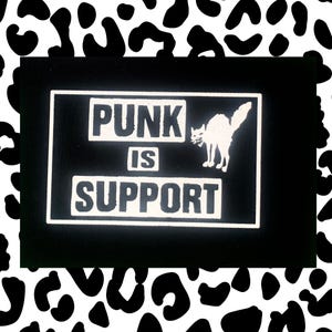 Könnte beinhalten: Schwarzer rechteckiger Aufnäher mit weißem Text „PUNK IS SUPPORT“. Eine weiße Katzen-Silhouette befindet sich rechts vom Text. Der Hintergrund ist schwarz und der Rand weiß. Der Hintergrund ist ein schwarz-weißes Dalmatiner-Muster.