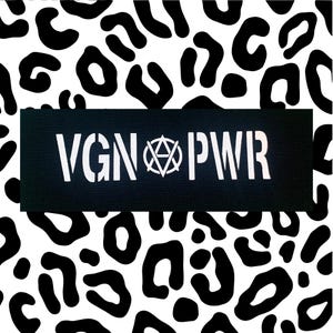 Könnte beinhalten: Ein schwarzes Rechteck mit dem weißen Text "VGN PWR" und einem zentralen Symbol, vor einem schwarz-weißen Leopardenmuster. Das Design ist auffällig und grafisch.