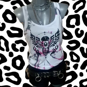 Könnte beinhalten: Ein weißes Tanktop mit einem Batik-Muster und einer schwarzen und weißen Totenkopfgrafik mit Flügeln. Das Tanktop hat ein rosa und graues Batik-Muster.