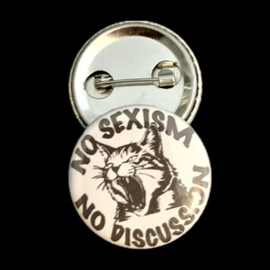 Könnte beinhalten: Ein runder Button mit dem Text "NO SEXISM NO DISCUSSION", der eine schwarz-weiße Illustration einer Katze umgibt. Der Button ist weiß mit schwarzem Text und einer schwarz-weißen Katzenillustration.
