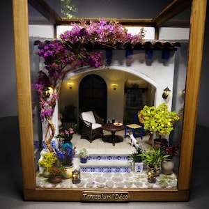 May include: A miniature diorama in a wooden frame titled Terraplum' Déco, featuring a white Mediterranean-style patio with blue and white tiled steps, a dark wicker chair, a small round table, potted plants, and vibrant purple bougainvillea.