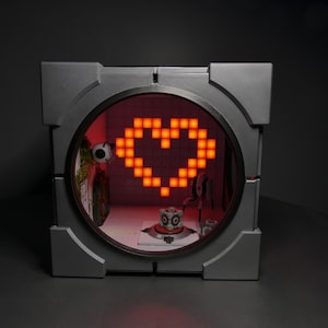 Könnte beinhalten: Eine graue, quadratische Leuchtbox mit einer kreisförmigen Öffnung, die ein pixeliges orangefarbenes Herz zeigt. Im Inneren der Box befinden sich Miniaturfiguren und Requisiten aus dem Videospiel Portal. Der Hintergrund ist dunkel.