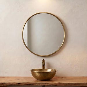 Peut inclure: Un miroir mural rond doté d'un cadre en métal doré fin, placé au-dessus d'une vasque à poser en laiton martelé. La vasque présente une finition métallique et est associée à un robinet doré assorti, créant un ensemble harmonieux pour la salle de bain.