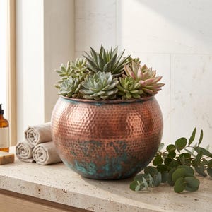 Peut inclure: Un pot de fleurs rond en cuivre martelé avec une finition patinée bleu-vert à la base, rempli d'une variété de plantes succulentes vertes et aux pointes roses. Le pot a une forme sphérique large et une surface texturée aux tons métalliques.