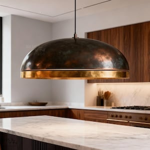 Puede incluir: Una lámpara colgante grande en forma de cúpula con acabado de cobre y una banda de latón. La luz cuelga de un cable negro en una cocina con encimera de mármol y armarios de madera.