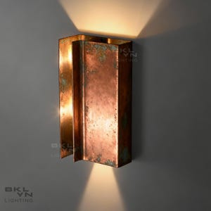 Peut inclure: Applique murale de couleur cuivre de forme rectangulaire. L'applique présente un aspect vieilli et patiné avec des touches de vert. Le luminaire est fixé sur un mur gris et émet une lueur chaude, avec le texte "BKLN LIGHTING".