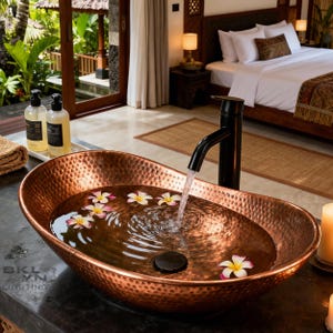 Peut inclure: Un évier de salle de bain en cuivre martelé rempli d'eau et de fleurs flottantes. Un robinet noir verse de l'eau dans l'évier. L'évier est dans une salle de bain bien éclairée avec un lit en arrière-plan.