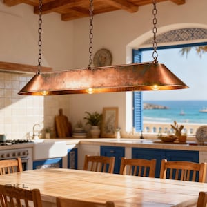 May include: A copper-colored, rectangular pendant light fixture with three exposed bulbs hangs from chains. The fixture is suspended above a wooden dining table in a kitchen with a view of the ocean. The fixture has a rustic, aged appearance.