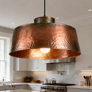 May include: A copper pendant light fixture with a hammered texture and a warm, reddish-brown hue. The light hangs from a black cord and illuminates a kitchen setting. The fixture has a wide, bowl-shaped shade and a small, round top.