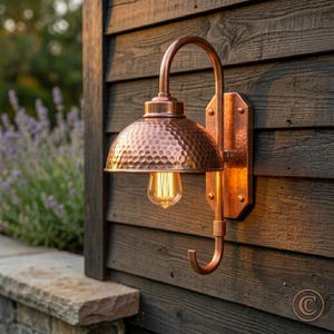 May include: A copper-colored outdoor wall-mounted light fixture. The light has a hammered dome-shaped shade and a curved arm. The light bulb is visible. The fixture is mounted on a wooden wall.