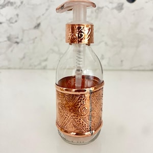 Peut inclure: Distributeur de savon en verre transparent avec une bande et une pompe en métal couleur cuivre. La bande métallique présente un motif en relief complexe. La pompe est en plastique transparent. Le distributeur est sur une surface blanche.