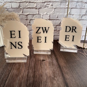 Könnte beinhalten: Drei Acryl-Tischnummernschilder mit hellbraunem Hintergrund und schwarzem Text. Die Nummern sind "Eins", "Zwei" und "Drei".