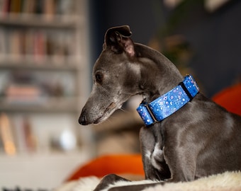 May include: A grey whippet dog wearing a blue and white patterned collar sits on a white furry surface. The dog is looking to the left of the frame.