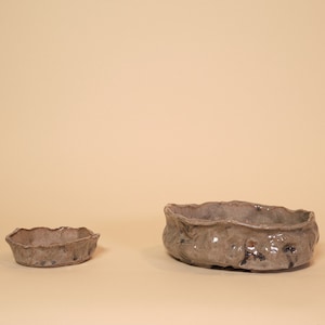Puede incluir: Dos macetas de bonsái de cerámica, una pequeña y otra grande, con un esmalte marrón claro y bordes irregulares. Las macetas están sobre un fondo beige claro.