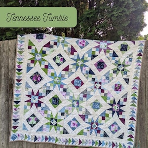 Peut inclure: Une courtepointe faite à la main avec un fond crème et un motif d'étoiles dans des tons de bleu, vert et violet. La courtepointe présente le texte "Tennessee Tumble" et "Scrap Basket Quilts". La courtepointe est exposée sur une clôture en bois.