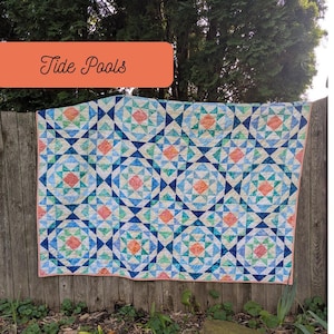 May include: A handmade quilt with a geometric pattern in shades of blue, green, and coral, hanging on a wooden fence. The quilt features the text "Tide Pools" in an orange rectangle. The words "Scrap Basket Quilts" are at the bottom.