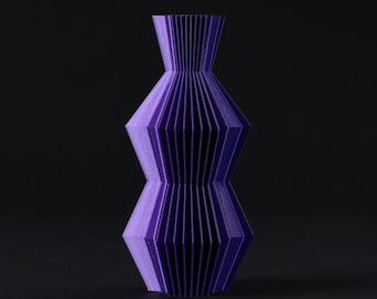 May include: A purple decorative vase with a geometric, concertina-like design. The vase has a wide base, a narrower middle section, and a flared top. The vase is made of a solid material and stands on a dark surface.