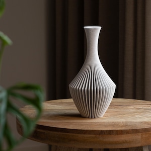 Op de afbeelding: Een witte, getextureerde vaas met een gegroefd ontwerp. De vaas heeft een brede basis die taps toeloopt naar een smalle hals en een uitlopende opening. Het staat op een ronde, houten tafel. De vaas is ongeveer 25 cm hoog.