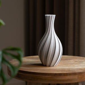 May include: A decorative vase with a striped pattern in shades of grey and white. The vase has a slender neck and a rounded body, sitting on a wooden table. The vase is a home decor item.
