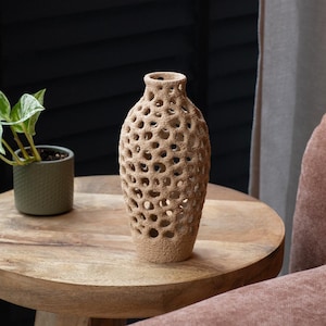 Puede incluir: Un jarrón beige texturizado con un diseño único de agujeros abiertos. El jarrón es cilíndrico con un cuello estrecho y está sobre una mesa redonda de madera. A la izquierda del jarrón se ve una pequeña planta en maceta.