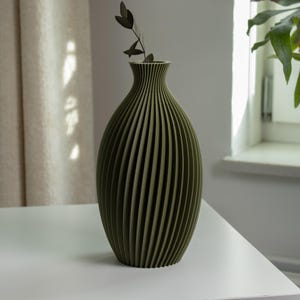 Könnte beinhalten: Olivgrüne Vase mit geripptem, vertikalem Design. Die Vase hat einen schlanken Hals und einen abgerundeten Körper. Ein kleiner Zweig getrockneter Blätter befindet sich in der Vase. Die Vase steht auf einem weißen Tisch.