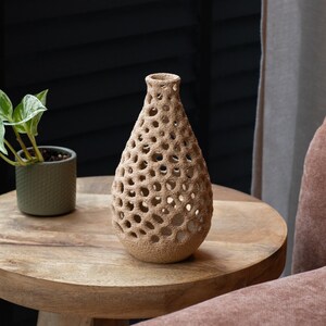 Può includere: Un vaso beige a forma di goccia con un design poroso strutturato. Il vaso decorativo è realizzato in un materiale di colore chiaro e poggia su un tavolo di legno. Una piccola pianta in vaso è visibile sullo sfondo.