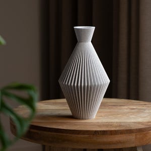 Puede incluir: Un jarrón gris geométrico con un diseño plisado texturizado. El jarrón tiene forma de diamante con un cuello estrecho y una abertura acampanada. Está sobre una mesa redonda de madera, mostrando una estética moderna y minimalista. El jarrón mide unos 30 cm de alto.