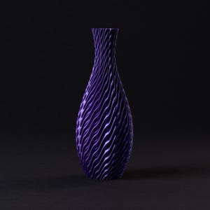 Puede incluir: Un jarrón alto y morado con un patrón texturizado en forma de olas. El jarrón tiene un cuello delgado y un cuerpo redondeado. El fondo es negro sólido, resaltando el diseño y el color únicos del jarrón.