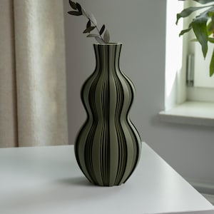 Può includere: Un vaso a righe verdi e nere con una forma curva unica. Il vaso contiene alcuni rametti di vegetazione. Il vaso è su un tavolo bianco, con una tenda e una finestra sullo sfondo.