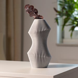 以下が含まれることがあります： モダンで幾何学的な花瓶。薄いグレーで、ユニークなプリーツデザインが特徴です。花瓶は細長い砂時計型で、乾燥した赤褐色の葉の小枝が入っています。花瓶は白い表面に置かれています。