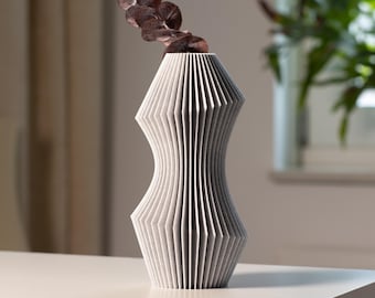 May include: A contemporary, geometric vase in light grey, with a distinctive pleated design. The vase has a slender, hourglass shape and contains a sprig of dried, reddish-brown leaves. The vase is displayed on a white surface.