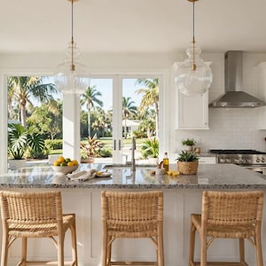 Puede incluir: Una isla de cocina con encimera de granito moteado cuenta con tres taburetes de ratán tejido. Sobre la isla cuelgan dos lámparas colgantes de cristal transparente. La cocina incluye armarios blancos y una campana extractora de acero.