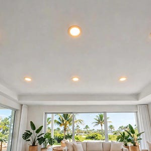 May include: A white ceiling featuring six circular recessed lights arranged in two rows. The lights emit a warm, soft glow, illuminating the interior space which includes large glass sliding doors and potted green plants.