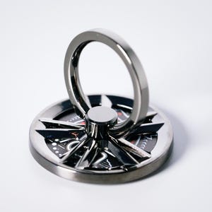 Puede incluir: Un soporte de anillo para teléfono plateado con un diseño de brújula. El anillo circular está en posición vertical, unido a una base redonda con un patrón de rosa de los vientos en negro y rojo. El soporte es de metal y está sobre una superficie blanca.