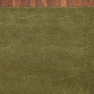 May include: A rectangular olive green rug with a soft, textured surface. The rug is placed on a wooden floor with a warm, brown tone. The corner of the rug is visible, showcasing its plush texture and color.