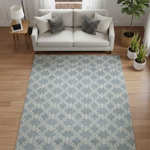 Op de afbeelding: Een rechthoekig vloerkleed met een lichtblauwe achtergrond en een herhalend wit vierpaspatroon. Het kleed ligt op een houten vloer in een woonkamer, met een witte bank, kussens en planten op de achtergrond.