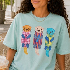 May include: A light mint green t-shirt featuring a graphic of three otters, each wearing a different coloured patterned shirt and holding a small fish. The otters are depicted with simple facial features, including X-shaped eyes.