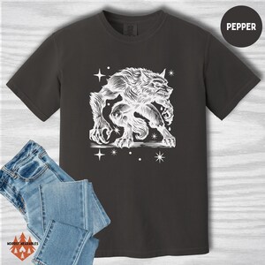 Flat lay of a pepper-colored Comfort Colors tee displaying a white linocut-style werewolf crouched in an aggressive pose, surrounded by cosmic stars. Styled with folded jeans. “PEPPER” color label at top right.
