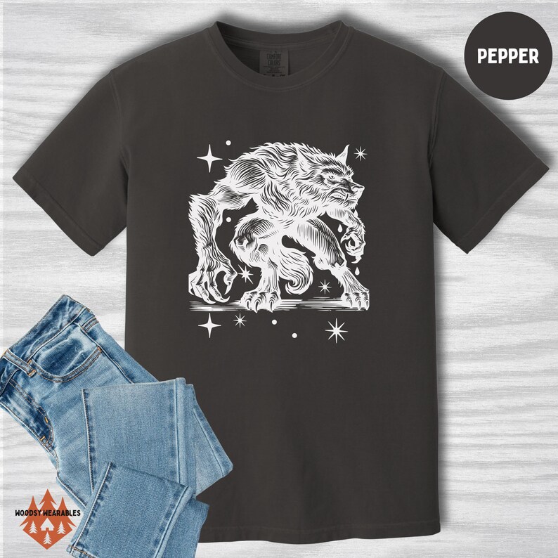 Flat lay of a pepper-colored Comfort Colors tee displaying a white linocut-style werewolf crouched in an aggressive pose, surrounded by cosmic stars. Styled with folded jeans. “PEPPER” color label at top right.