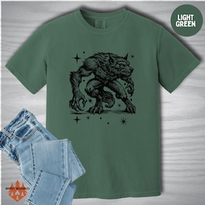 Flat lay of a Comfort Colors shirt in light green featuring a black werewolf design in linocut style. The werewolf crouches surrounded by stars, printed alongside a folded pair of jeans. “LIGHT GREEN” label in the upper right.